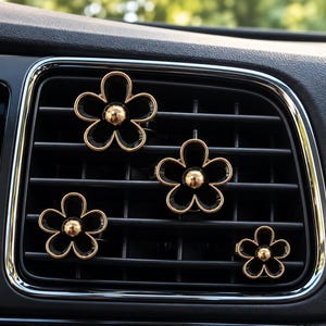 Könnte beinhalten: Vier blumenförmige Auto-Lüftungsclips mit schwarzen Blütenblättern, goldfarbenen Metallrändern und einer goldenen Kugel in der Mitte. Diese dekorativen Accessoires sind an den horizontalen Lamellen einer schwarzen Lüftungsdüse befestigt.