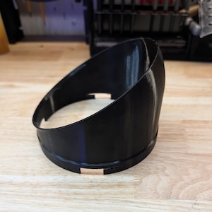 Puede incluir: Una cubierta cilíndrica de plástico negro impresa en 3D con un borde superior inclinado. El componente presenta una base circular con pequeños recortes rectangulares a lo largo del borde inferior, diseñados para montaje o alineación.