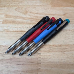 May include: Four pocket screwdrivers with metallic tips and textured barrels in black, red, blue, and black. Each tool features a pocket clip and a knurled end cap. The screwdrivers are arranged diagonally, showcasing their distinct colored bodies and silver-toned metal components.