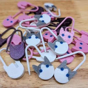 May include: A collection of plastic stitch markers shaped like cat heads in various colors including pink, white, gray, and dark purple. Each marker features a U-shaped clasp mechanism with small blue dots representing eyes on the cat-shaped heads.