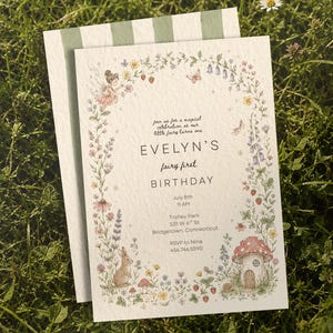 May include: A white rectangular birthday invitation card featuring a floral border with a fairy, a rabbit, and a mushroom house. The text reads: Join us for a magical celebration as our little fairy turns one, EVELYN'S fairy first BIRTHDAY, July 8th, 11 AM, Trolley Park, 501 W 6th St, Bridgetown, Connecticut, RSVP to Nina, 456.746.5590.