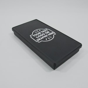 May include: A rectangular black box with a flat lid featuring a white circular graphic print in the center. The print contains the text "YOUR TEXT LOGO OR GRAPHIC HERE" surrounded by a decorative border with small radiating lines at the top and bottom.