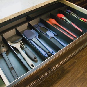 May include: A kitchen drawer organizer with black dividers containing various cooking utensils including a black slotted turner, a grey spatula, blue silicone spatulas, a red spoon, and metal tongs with red silicone tips arranged in separate slots.
