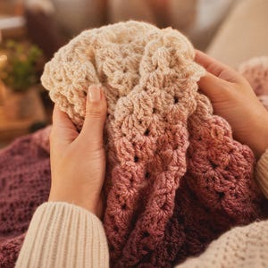 May include: A crocheted blanket featuring a gradient color transition from cream to light pink, dusty rose, and deep plum. The textile displays a textured, repeating stitch pattern throughout, showcasing a soft, yarn-based construction.