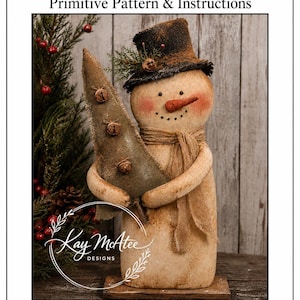 Pode incluir: Uma figura de boneco de neve de tecido segurando uma árvore de tecido verde, com uma cartola preta e um cachecol bege. O texto diz Marvin's Tree Primitive Pattern & Instructions, Kay McAtee Designs e 24" TALL. O boneco tem nariz de cenoura e bochechas rosadas.