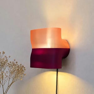 Könnte beinhalten: Eine Wandleuchte mit zweifarbigem Design, bestehend aus einem durchscheinenden pfirsichfarbenen oberen Teil und einem massiven dunkelvioletten unteren Teil. Die Leuchte strahlt warmes Licht nach oben und unten ab, mit einem schwarzen Kabel.