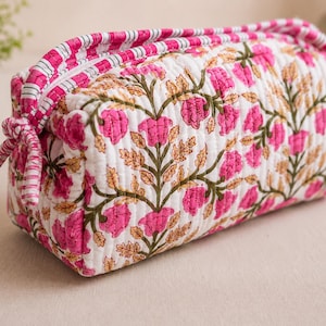 May include: A rectangular quilted fabric pouch featuring a white base with a repeating pattern of pink flowers and green stems. The zipper closure is accented with a pink and white striped fabric trim and a knotted fabric pull on the side.