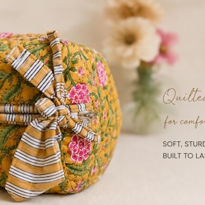 May include: A quilted mustard yellow pouch with a floral pattern in pink and green. It features a zipper closure with a striped fabric bow tie. The text on the right reads: Quilted for comfort, SOFT, STURDY & BUILT TO LAST.