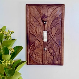 May include: A rectangular dark brown wooden light switch plate featuring an intricate carved floral pattern. The plate is mounted on a wall and includes a central white toggle switch secured by two small silver-coloured metal screws.