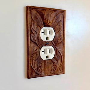 May include: A rectangular wooden wall plate cover featuring intricate carved leaf patterns surrounding a standard white dual electrical outlet. The wood has a medium brown finish with visible grain, and the plate is mounted flush against a plain wall.