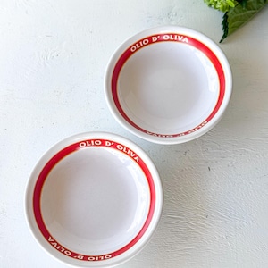 May include: Two white ceramic bowls featuring a red circular band along the inner rim. The text OLIO D' OLIVA is printed in white capital letters within the red band on both bowls. The bowls have a smooth, glossy finish and a simple round shape.
