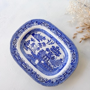 May include: An oval ceramic serving platter featuring a blue and white willow pattern. The center depicts a landscape with a pagoda, willow trees, and birds, while the wide rim is decorated with a geometric border and floral motifs.