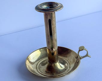 May include: A brass chamberstick candle holder featuring a circular drip pan base, a central cylindrical shaft with a vertical slot and an adjustable sliding thumb piece, and a small ring handle attached to the side of the base for carrying.