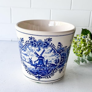 May include: A ceramic planter with a white base and blue decorative patterns. The front features an oval frame containing a detailed blue and white landscape illustration of a windmill. The rim and base are accented with thin blue circular lines.