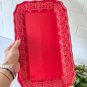 May include: A rectangular serving tray in a vibrant red color featuring a smooth, flat center and a raised, intricate border with a continuous, ornate, swirling filigree pattern along the entire edge of the tray, which has a wavy, scalloped rim.