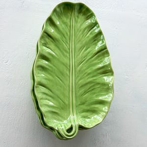 May include: A stack of three ceramic serving platters shaped like large, elongated green leaves. Each platter features a glossy finish, a prominent central vein, and textured ridges that mimic the natural appearance of a tropical plant leaf.