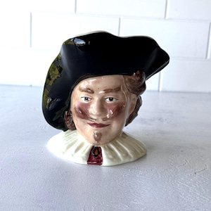 May include: A ceramic character jug depicting a person wearing a large black tricorn hat. The figure has brown hair, a mustache, and a goatee, and is dressed in a white ruffled collar with a small dark red accent at the center of the neckline.