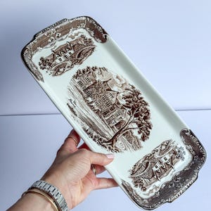 May include: A rectangular ceramic serving tray with a cream-colored base and brown transferware illustrations. The design features three distinct landscape scenes of buildings and trees, with decorative floral borders along the edges.