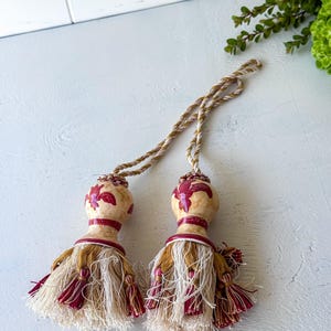 May include: A pair of decorative curtain tiebacks featuring cream-colored ceramic finials with a red floral pattern. Each finial is attached to a twisted beige and white cord, ending in a full tassel composed of cream, gold, and deep red threads.