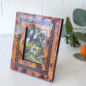 May include: A rectangular picture frame featuring a polished burl wood finish with a dark brown, intricate geometric inlay pattern bordering the central glass pane. The frame has a glossy, smooth surface and a vertical orientation.