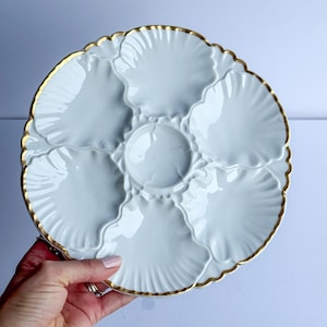 May include: A white ceramic oyster plate featuring six shell-shaped indentations arranged around a central circular well. The plate has a scalloped edge and is finished with a thin gold trim along the entire outer rim and the inner dividers.