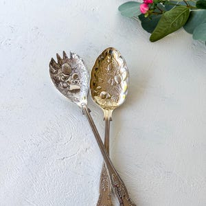 May include: A pair of silver-toned metal salad serving utensils featuring an embossed fruit and foliage design on the bowls and handles. One utensil is a solid spoon, while the other is a slotted spoon with three tines at the top edge.