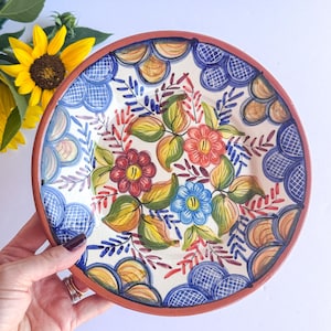 May include: A round ceramic bowl with a terracotta rim featuring a hand-painted floral design in red, blue, and green. The interior pattern includes stylized flowers and leaves surrounded by a border of blue and tan scalloped shapes.