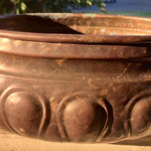 May include: A round copper pot featuring a wide opening, a smooth upper rim, and a lower section decorated with a series of embossed, rounded vertical panels. The vessel includes two curved metal handles attached to opposite sides of the body.