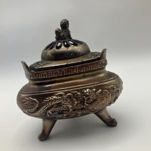 May include: A bronze-toned metal incense burner featuring three legs and a removable lid. The body is decorated with embossed dragon motifs, while the lid includes a small sculpted creature finial and cut-out ventilation holes.