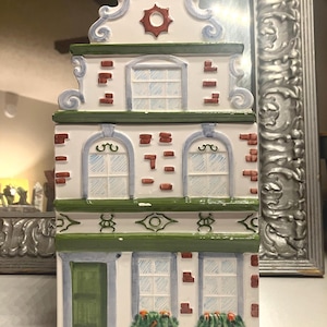 May include: A ceramic canister shaped like a tall, narrow Dutch canal house. The white facade features green trim, blue window outlines, red brick accents, and a green door. The roof is stepped with a central red circular emblem and blue detailing.