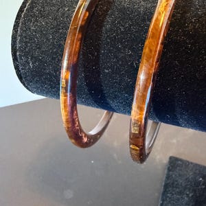 May include: Two circular bangle bracelets featuring a translucent amber-colored resin material with embedded gold-toned metallic flakes. The bracelets have a smooth, polished finish and a rounded profile, showcasing a warm, variegated brown and gold pattern.