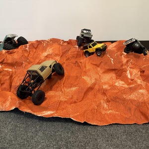 Op de afbeelding: Vijf miniatuur op afstand bestuurbare terreinwagens in diverse kleuren, waaronder beige, geel en groen, staan op een gestructureerd, gekreukt oranjebruin papieren oppervlak dat is ontworpen om op een rotsachtig woestijnterrein te lijken.