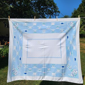 May include: A rectangular white tablecloth featuring a wide light blue border decorated with a repeating pattern of various teapots, teacups, and saucers. The central white panel is adorned with a subtle, light blue polka dot pattern throughout.