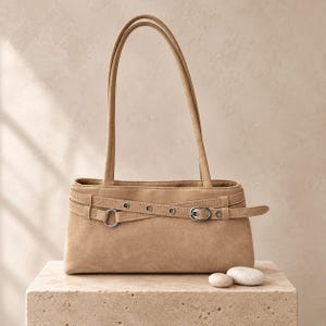 May include: A tan shoulder bag with a rectangular silhouette and two long handles. The exterior features a decorative horizontal strap across the front, accented with silver-toned metal eyelets, a circular ring, and a small adjustable buckle.