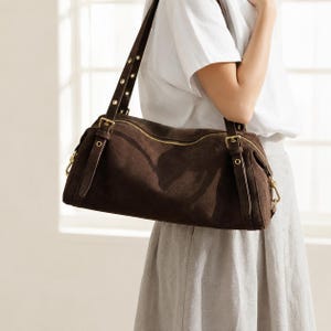 May include: A dark brown suede shoulder bag featuring a horizontal zipper closure, adjustable straps with metal buckles, and metallic eyelet details. The bag has a structured, elongated cylindrical shape with visible stitching along the edges.