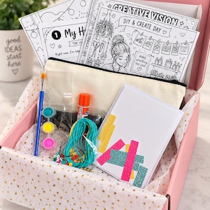 May include: A pink gift box containing a DIY craft kit with colouring sheets labelled Creative Vision and My H, a beige canvas pencil case, a blue paintbrush, three small paint pots, a glue stick, turquoise cord, beads, and a white card with glittery strips.