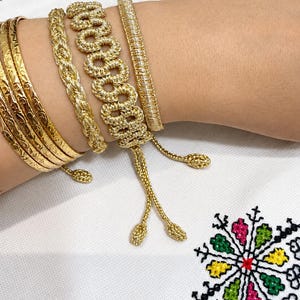 May include: A collection of gold-toned bracelets including three thin bangles with etched patterns, a braided rope-style bracelet, a wide woven link bracelet, and a smooth cord bracelet, all featuring dangling gold-toned end pieces with small knots.