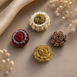 May include: Four round decorative buttons featuring intricate woven cord designs. One button is cream with a metallic olive center, one is deep red with a silver border, one is metallic gold, and one is a dark bronze brown with a floral knot pattern.