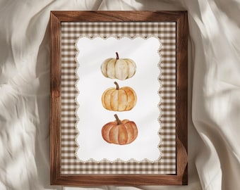May include: A framed wall art print featuring three watercolor pumpkins stacked vertically in the center. The background is a brown and white gingham pattern with a white scalloped border surrounding the pumpkins, all encased in a dark wood frame.