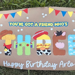May include: A brown paper sign featuring the text YOU'VE GOT A FRIEND WHO'S THREE and Happy Birthday Arlo. The word THREE is decorated with patterns inspired by cartoon characters, including a cowboy hat, space suit, and cow print details.