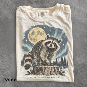 Puede incluir: Una camiseta de color marfil con un gráfico de un mapache de pie sobre un tocón de árbol. El fondo muestra una luna llena, un cielo estrellado y auroras boreales sobre un bosque. El texto "life is good in the woods" está impreso en la parte inferior.