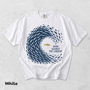 Könnte beinhalten: Ein weißes kurzärmeliges T-Shirt mit einer großen kreisförmigen Grafik aus dunkelblauen Fischen, die in einem Wellenmuster schwimmen. In der Mitte schwimmt ein einzelner goldener Fisch in die entgegengesetzte Richtung, neben dem Text "SWIM AGAINST THE CURRENT".