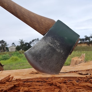Peut inclure: Une tête de hache en métal avec un manche en bois plantée dans un morceau de bois. La tête de la hache présente une section supérieure peinte en vert foncé et une lame en acier patiné avec le texte gravé CRAFTSMAN 4 visible sur le métal.