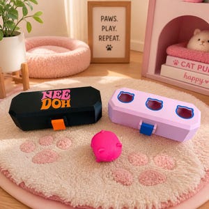 May include: A black rectangular storage case with the text NEE DOH in pink and orange, a light purple rectangular case with three cat-shaped cutouts, and a small bright pink cat-shaped toy figure resting on a textured surface.