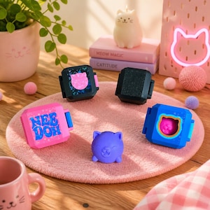 May include: Three square Nee Doh fidget boxes in pink, black, and blue, alongside a small purple cat-shaped figure. The pink box features the text Nee Doh in blue letters. These items are arranged on a round pink mat with a wooden surface visible.