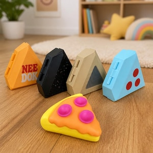 May include: A collection of five triangular fidget toys. Four are upright, including an orange one with the text NEE DOK, a black one with stars, a beige one, and a light blue one with red dots. A yellow and orange pizza-shaped fidget toy lies flat.