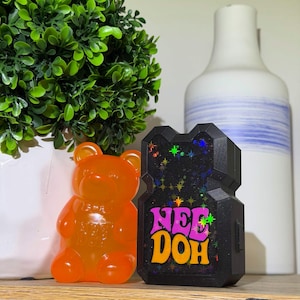 May include: A translucent orange bear-shaped stress toy with the text Nee Doh embossed on its chest, positioned next to a black rectangular stress toy featuring the text Nee Doh in pink and orange letters with holographic star accents.