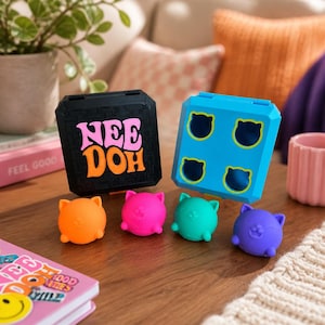May include: A black square case with the text NEE DOH in pink and orange, a blue square case with four cat-shaped cutouts, and four small cat-shaped squeeze toys in orange, pink, teal, and purple arranged in a row on a wooden surface.
