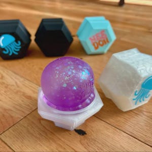 May include: A collection of five hexagonal fidget toys. Two are black with a blue jellyfish graphic, one is light blue with orange text reading NEE DOH, one is white with a blue jellyfish graphic, and one is a purple glitter sphere on a white base.