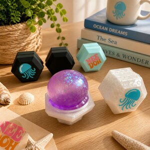 May include: A collection of Nee Doh sensory toys including a purple glittery sphere on a white hexagonal base, a white hexagonal container with a blue jellyfish graphic, a light blue container with Nee Doh text, and two black hexagonal containers.